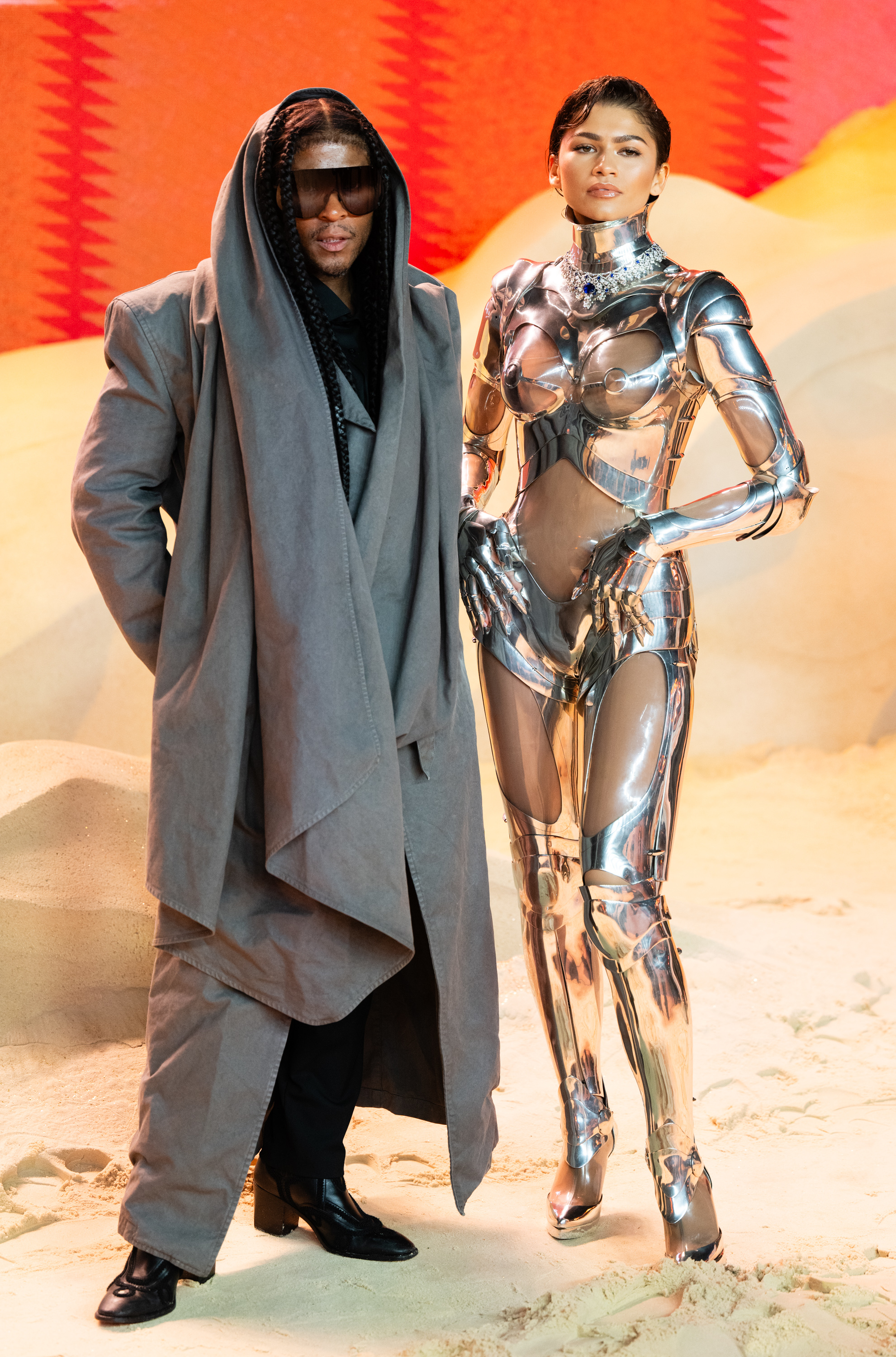 Two individuals posing, one in a metallic bodysuit and the other in an oversized gray coat