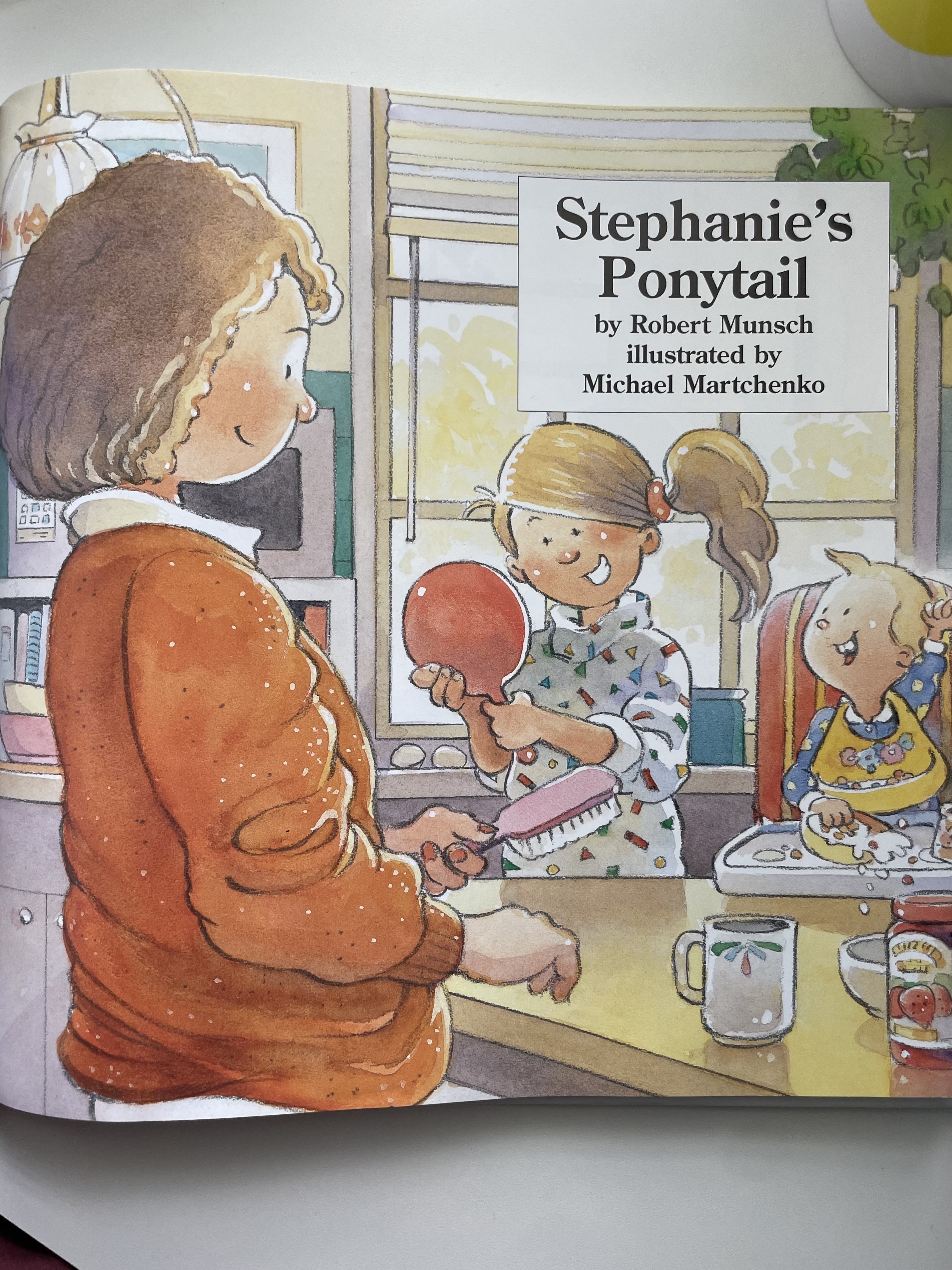 Illustration of a book cover titled &quot;Stephanie&#x27;s Ponytail&quot; with author names Robert Munsch and Michael Martchenko. Shows a girl with a ponytail facing a mirror held by an adult