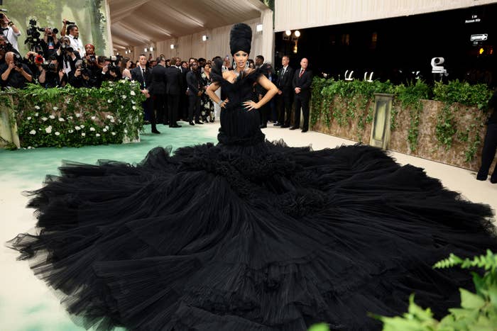 Person in an extravagant black gown with layers and headpiece on a carpet, photographers in the background