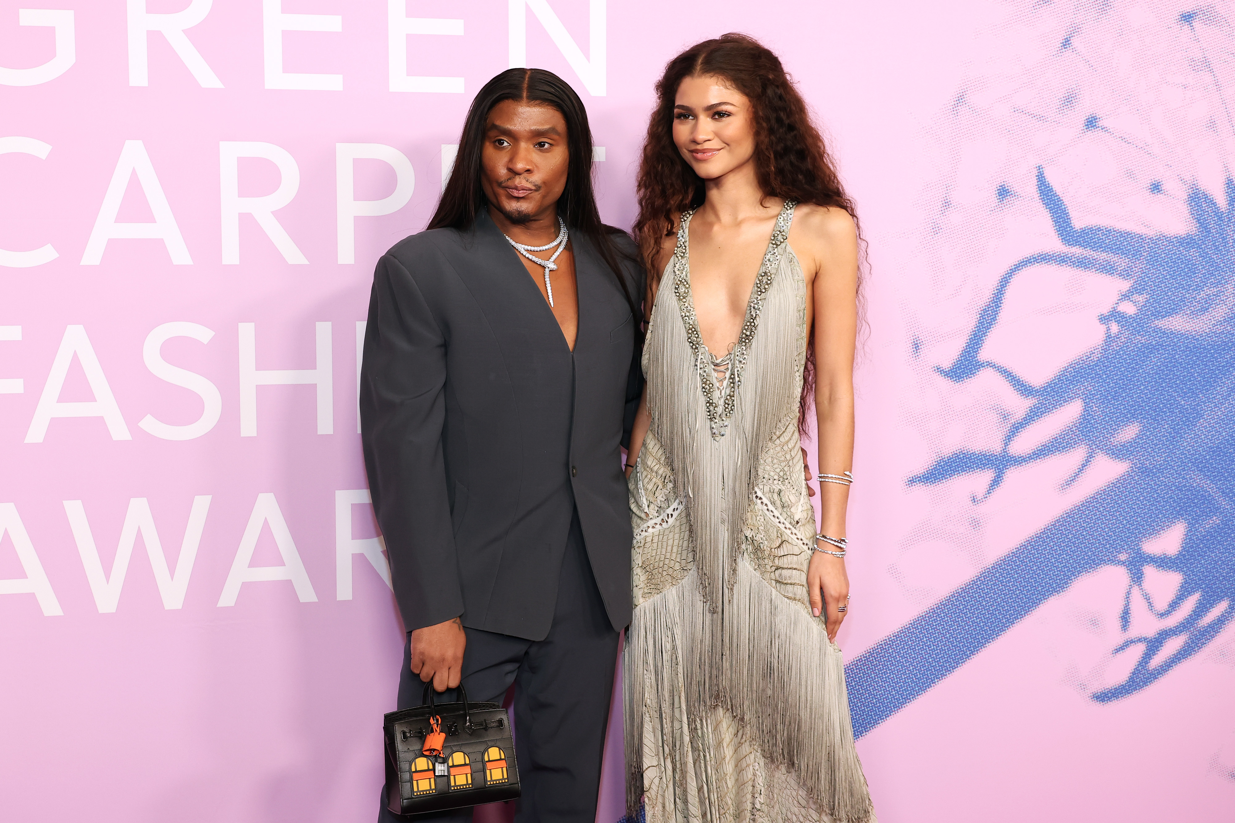 Two individuals posing at an event, one in a gray suit and holding a clutch, the other in a fringe-detailed dress