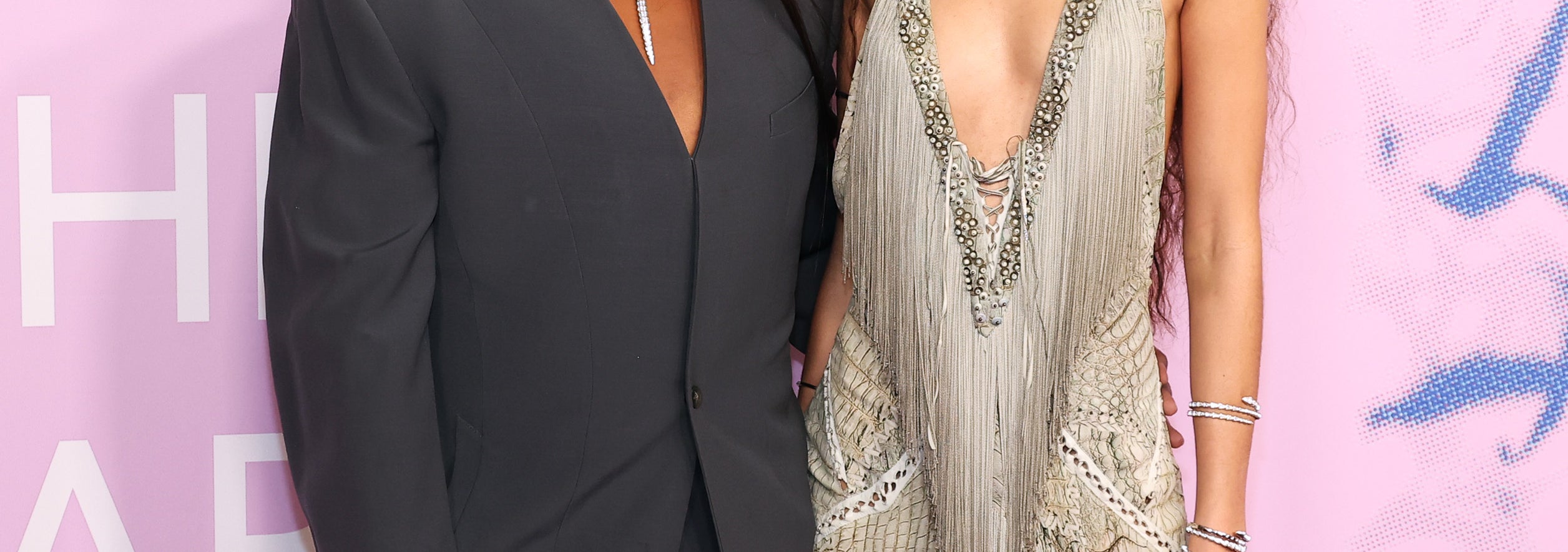 Two individuals posing at an event, one in a gray suit and holding a clutch, the other in a fringe-detailed dress