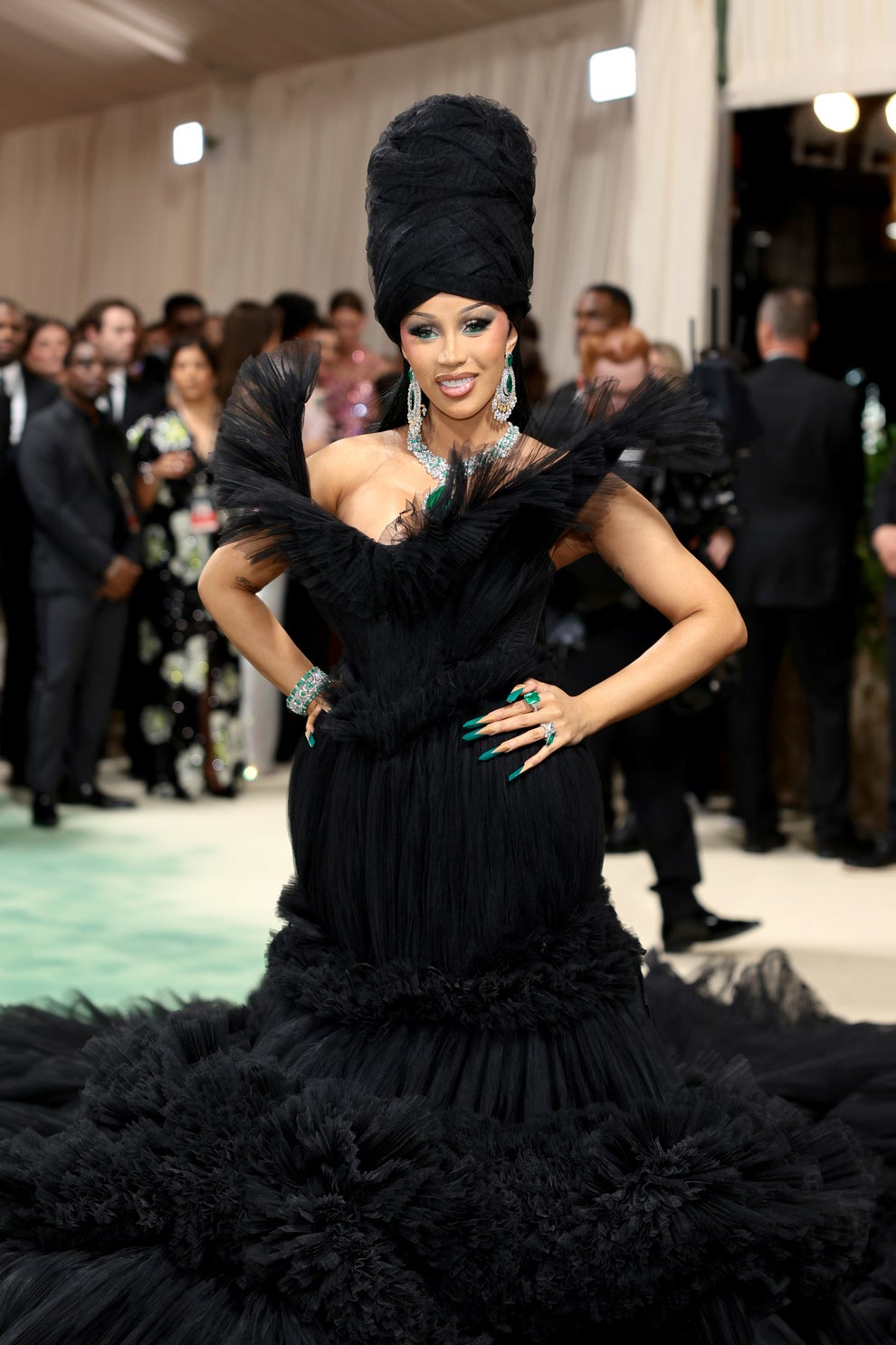 Person in an elaborate black dress and tall headpiece posing on a carpet with onlookers behind