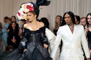 Two celebrities at an event; one in a large black gown with a floral headpiece, the other in an embellished white suit