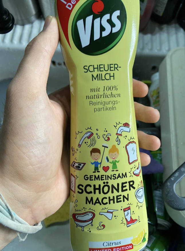 A hand holding a bottle of Viss cleaning product with whimsical cartoon characters and German text promoting communal cleaning