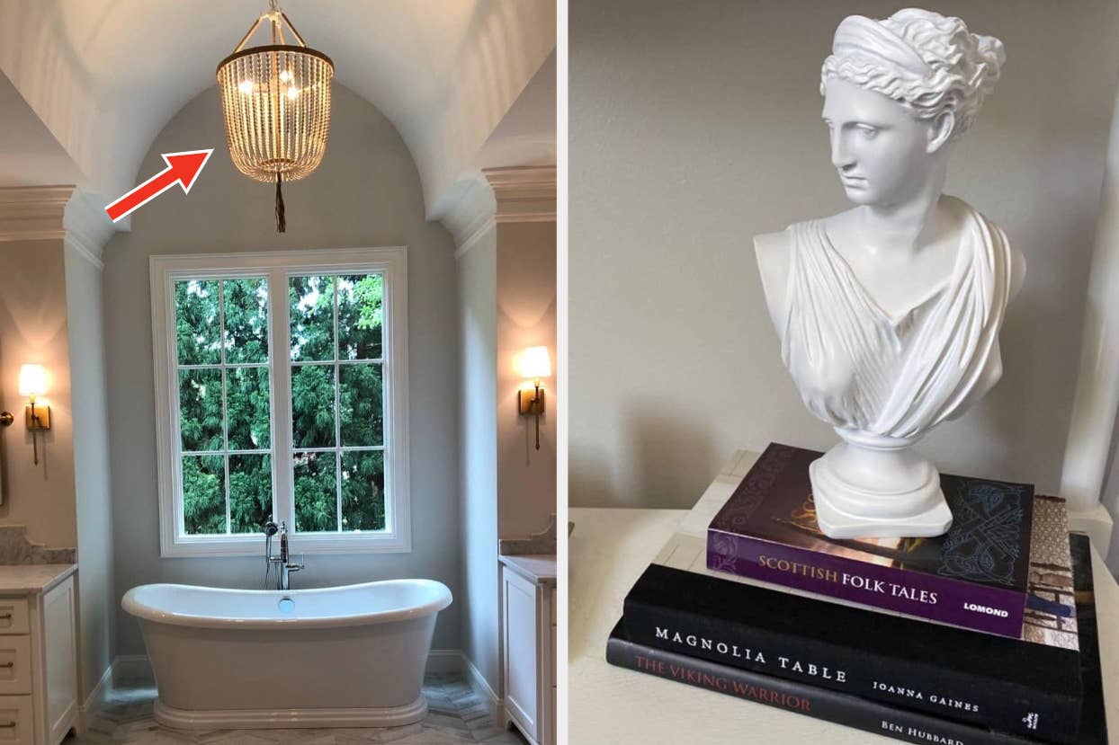 a freestanding bathtub by a window with a chandelier and a statue on books