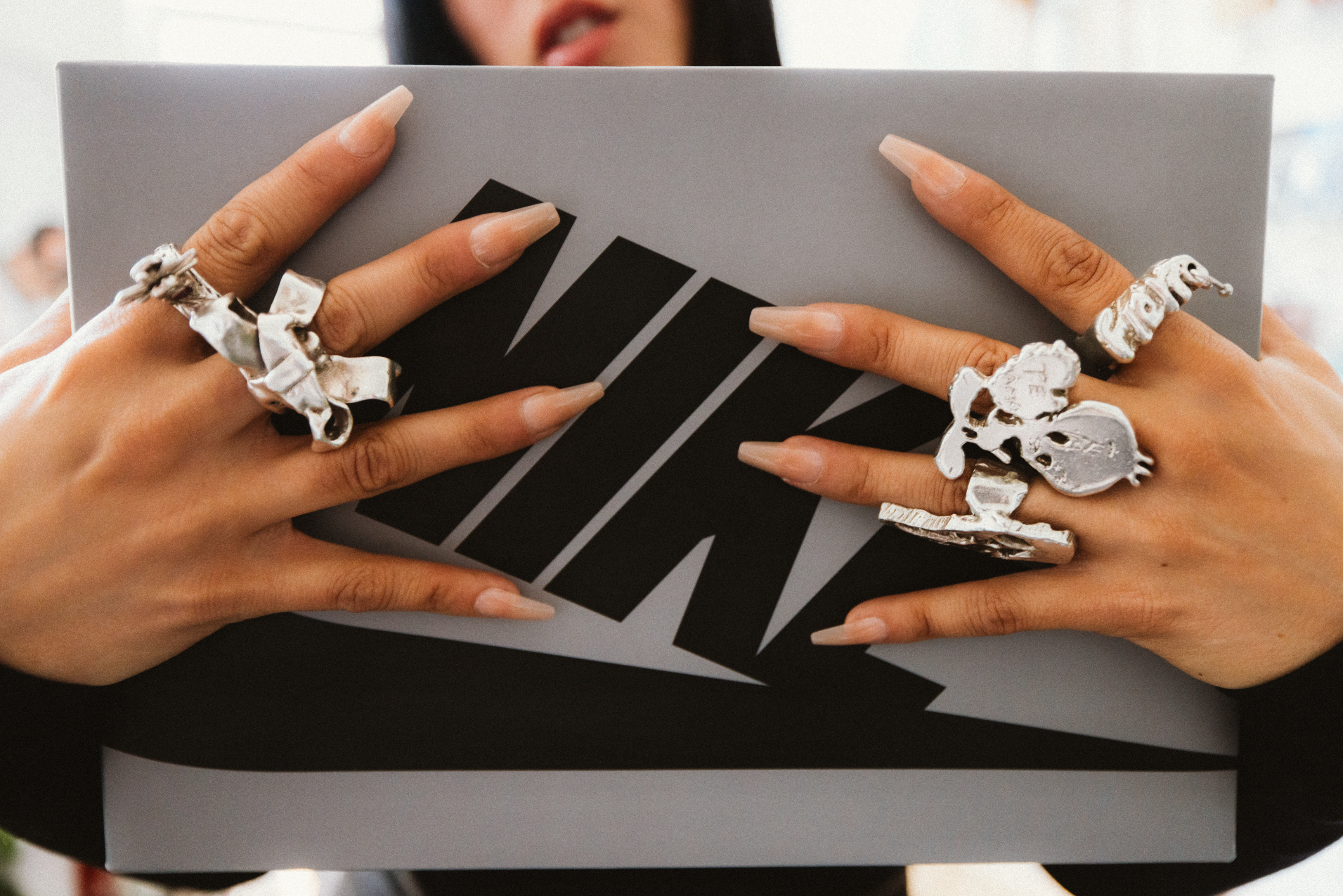 Close-up of hands holding a sneaker box with distinctive logo, adorned with silver rings including skull designs