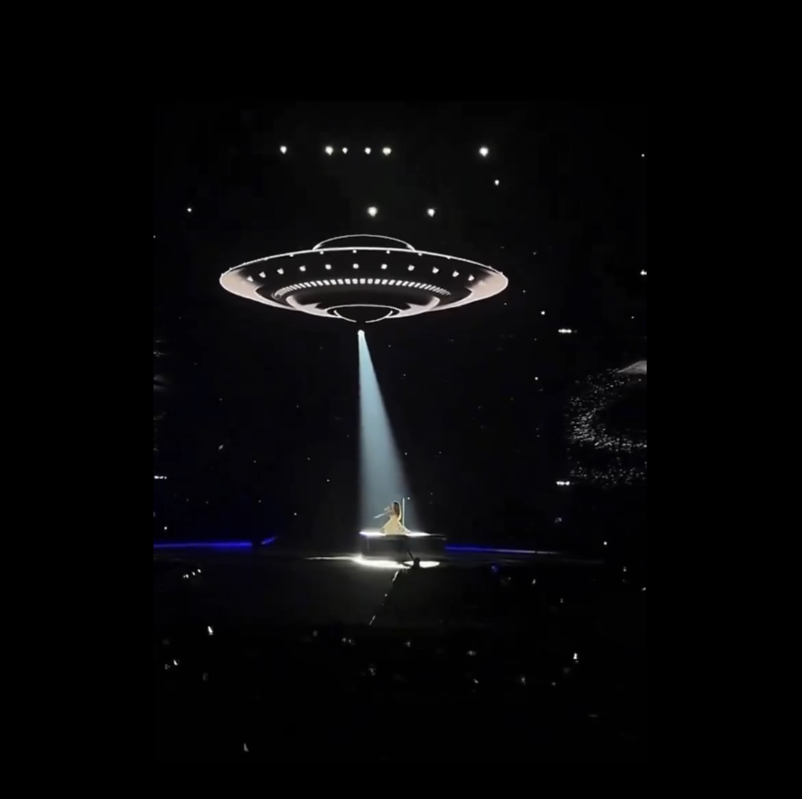 A performer plays piano under a large, illuminated ring on a darkened concert stage