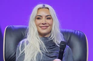 Kim Kardashian seated on stage, smiling, wearing a draped top and thigh-high boots