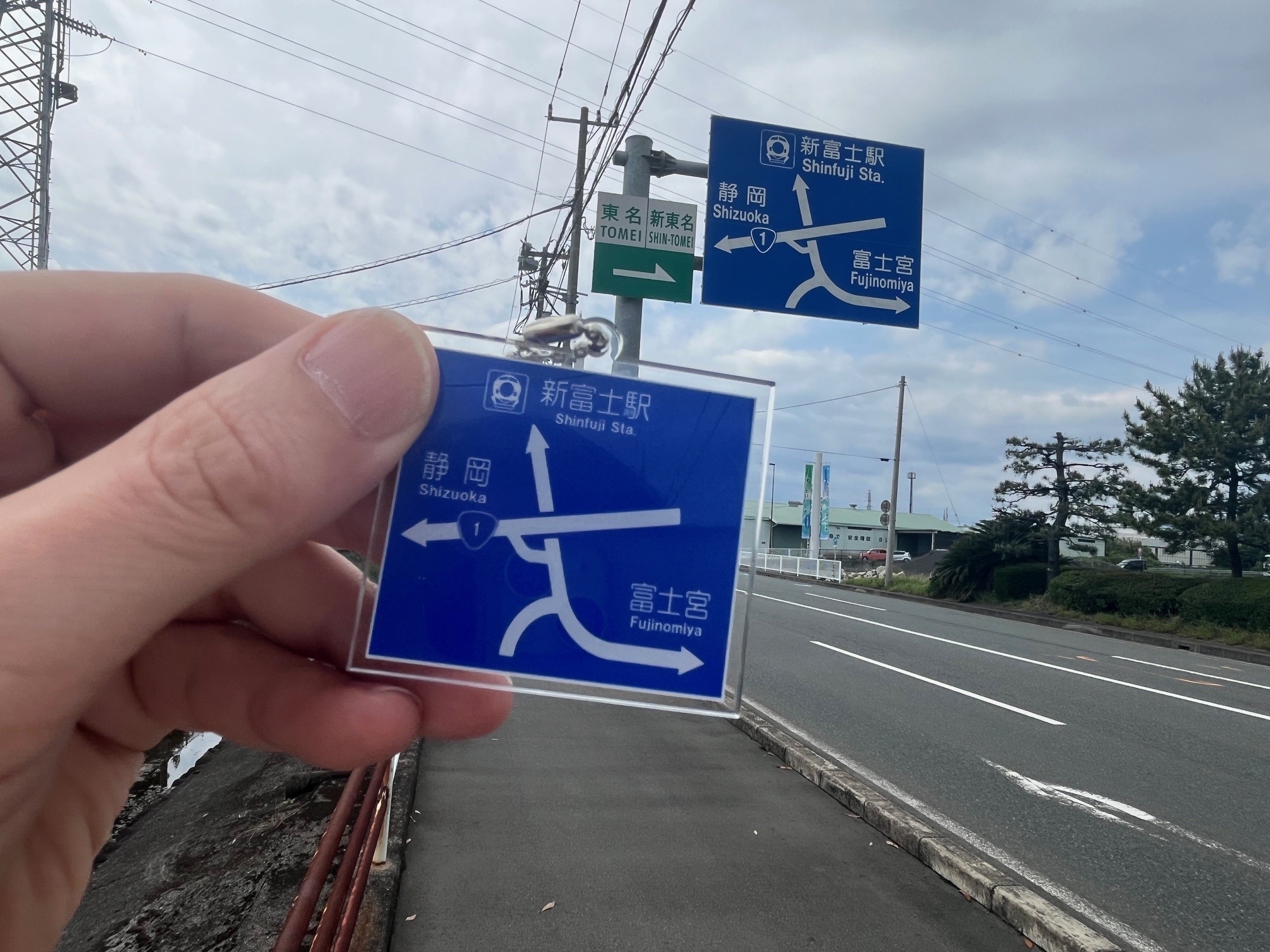 手前には道路標識を指し示すキーホルダーがあり、背景には同じ標識が実際に設置されている。