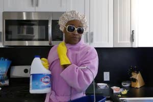 Person poses with cleaning supplies wearing a shower cap, sunglasses, gloves, and a bathrobe