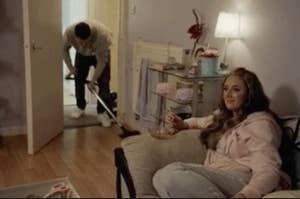 Woman sitting relaxed with a drink, smiling at a man vacuuming the room