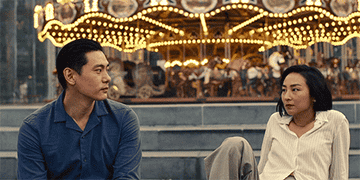 Two people are sitting and conversing in front of a carousel, expressing thoughtful emotions