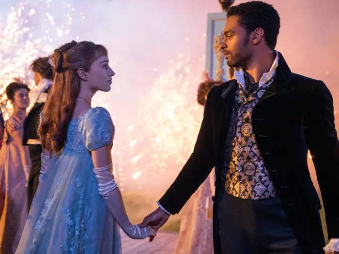 Dos personajes de "Bridgerton", protagonista femenina en vestido de época y protagonista masculino en atuendo formal, se toman de las manos