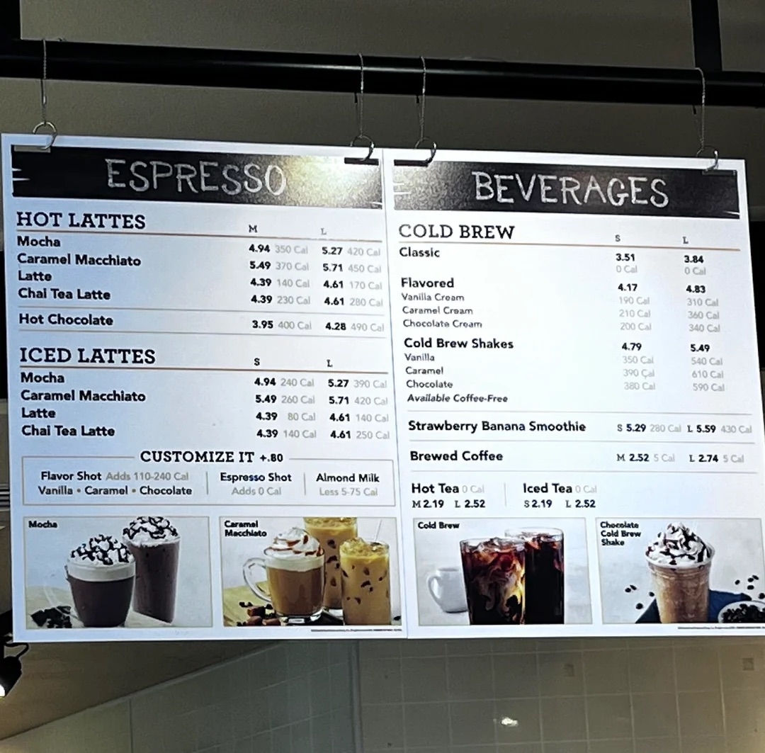 Menu board displaying a variety of hot lattes, iced lattes, cold brews, and tea options with prices at a coffee shop