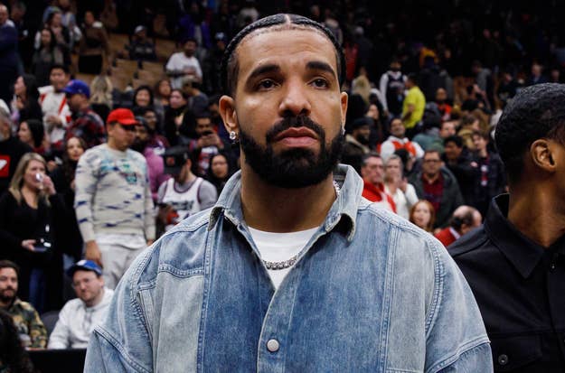 Drake in a denim jacket at a basketball game, looking pensive with crowds in the background