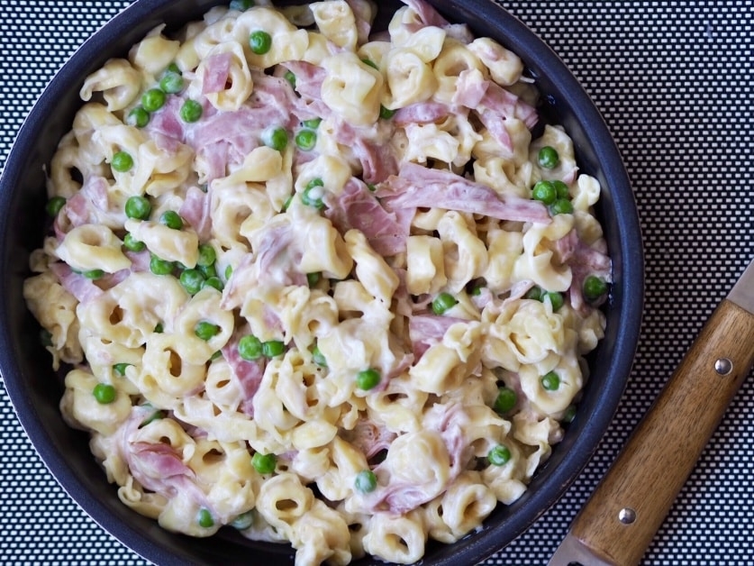 A skillet with creamy pasta, peas, and ham pieces, next to a wooden handle