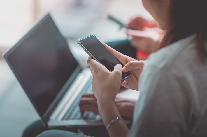 スマートフォンを操作する女性と、前面に見えるノートパソコン。