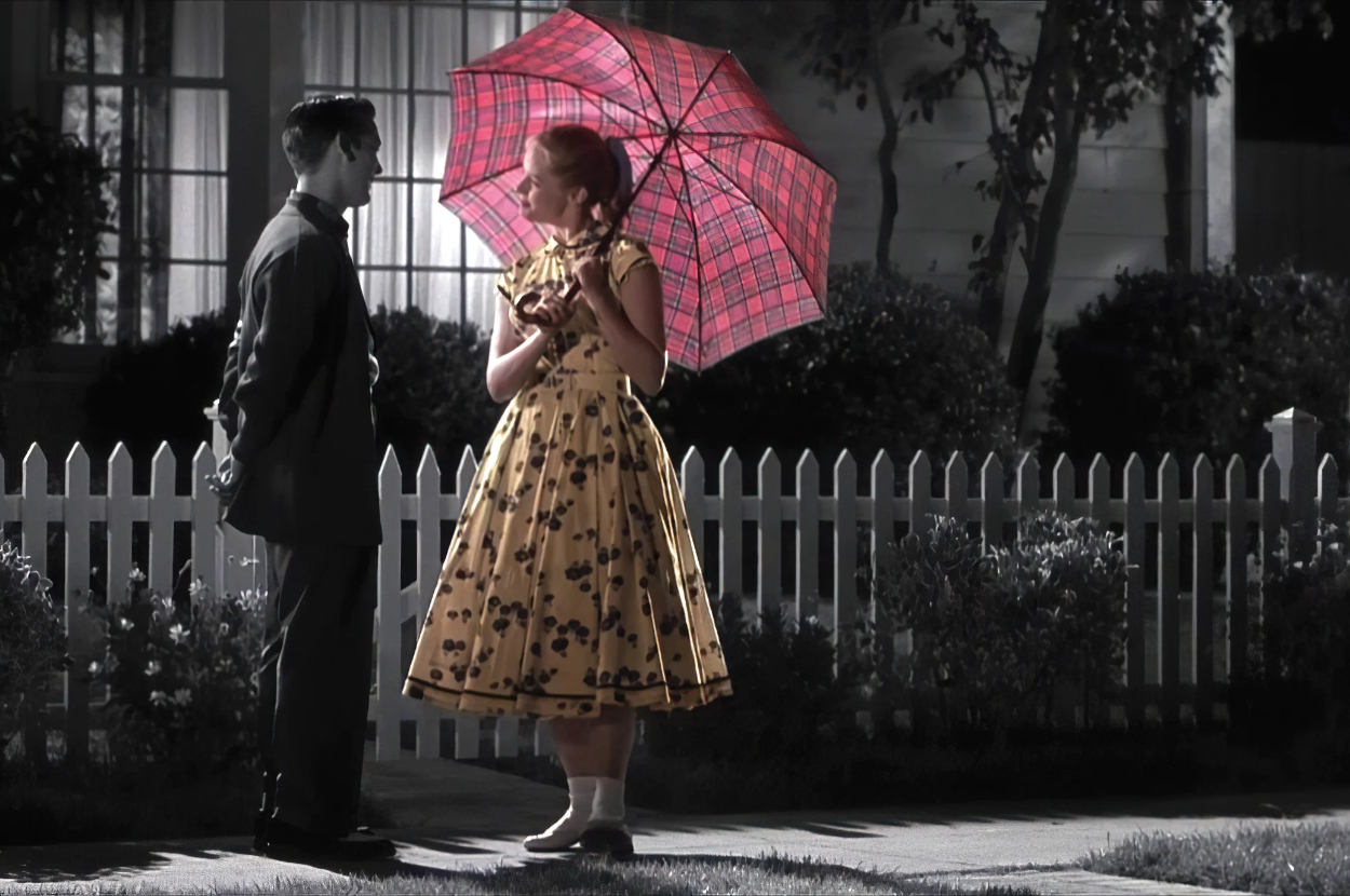 Two actors in vintage attire with a woman holding a patterned umbrella stand by a picket fence, reminiscent of a classic TV show scene