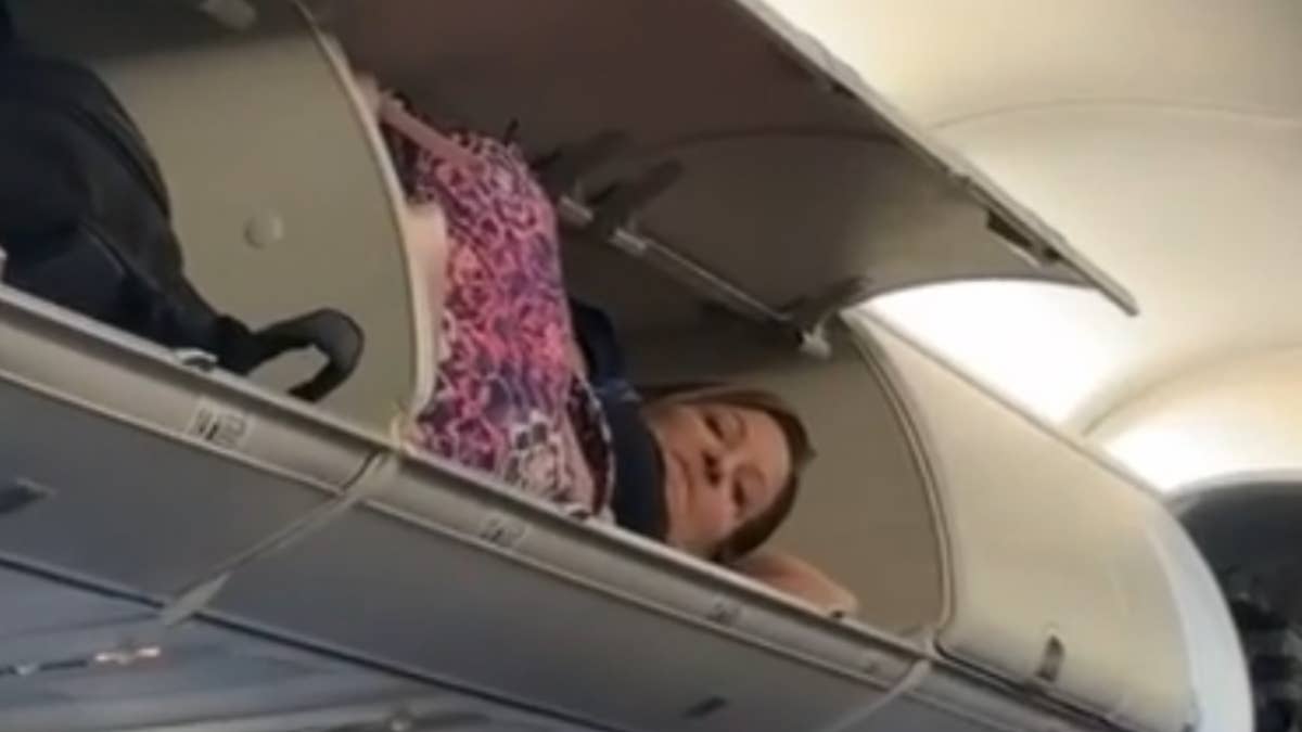 The woman was lying down with a piece of luggage as passengers walked to their seats.