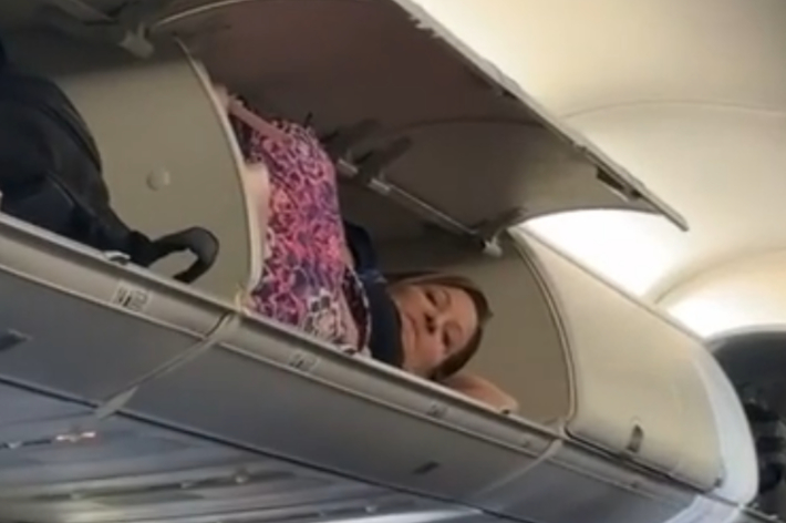Person laying in an overhead luggage compartment on an airplane