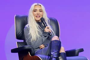Kim Kardashian seated on stage in a chair speaking into a microphone wearing a blazer and high boots