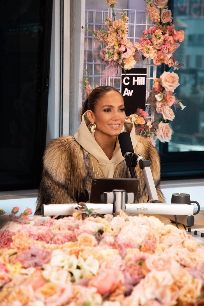 Jennifer Lopez wearing a fur coat, sitting at a microphone surrounded by flowers