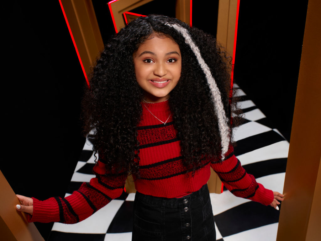 Young girl smiling, styled in a striped sweater and denim skirt, stands in a graphic room with a checkered floor and angular lines