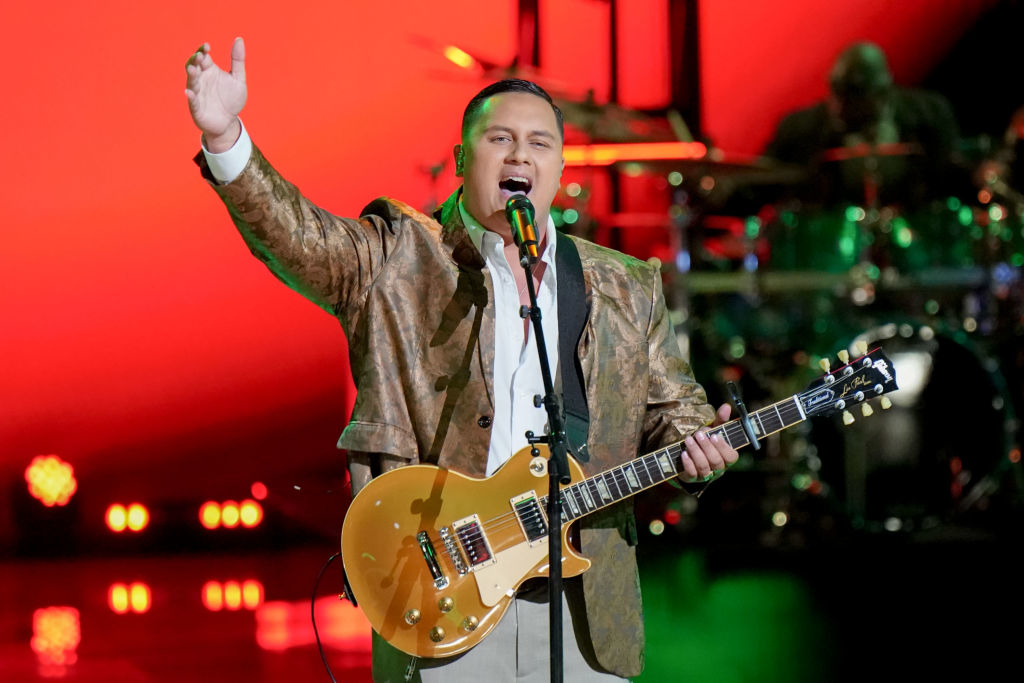 Musician in a patterned jacket, performing with a guitar on stage with band and red backdrop