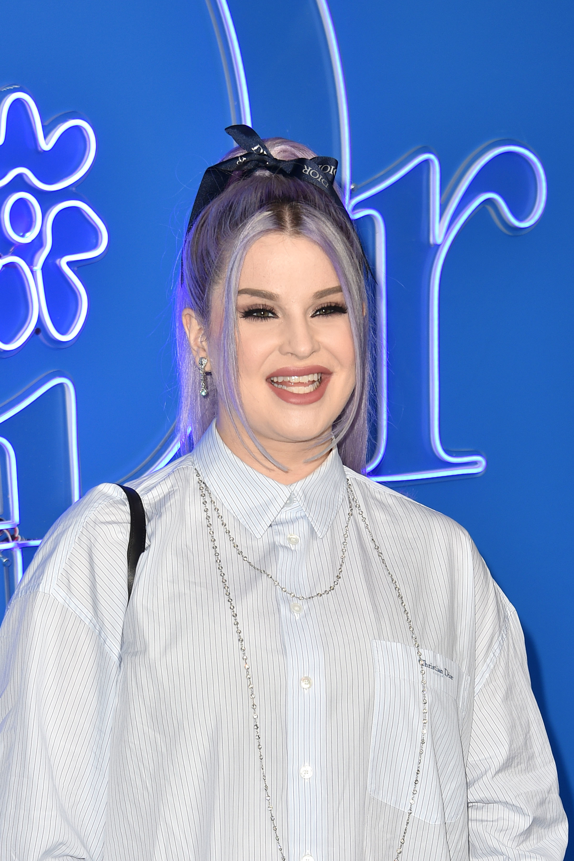 Kelly Osbourne wearing a striped shirt and bow in her hair, smiling at an event with a neon backdrop