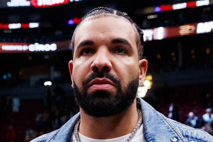 Drake wearing a denim jacket at a sports event, looking into the camera
