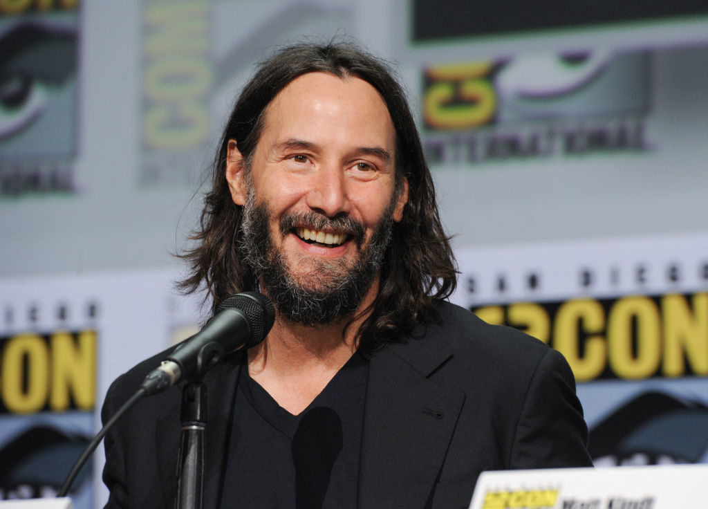 Keanu Reeves smiling, seated at Comic-Con panel, wearing a black suit without a tie