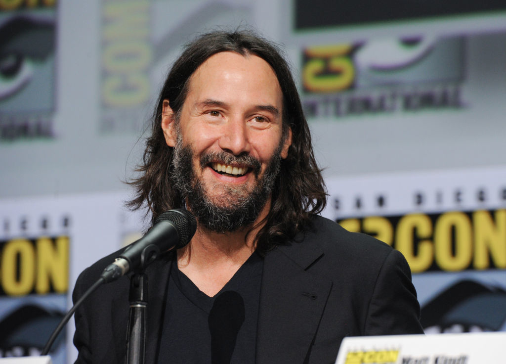 Keanu Reeves smiling, seated at Comic-Con panel, wearing a black suit without a tie