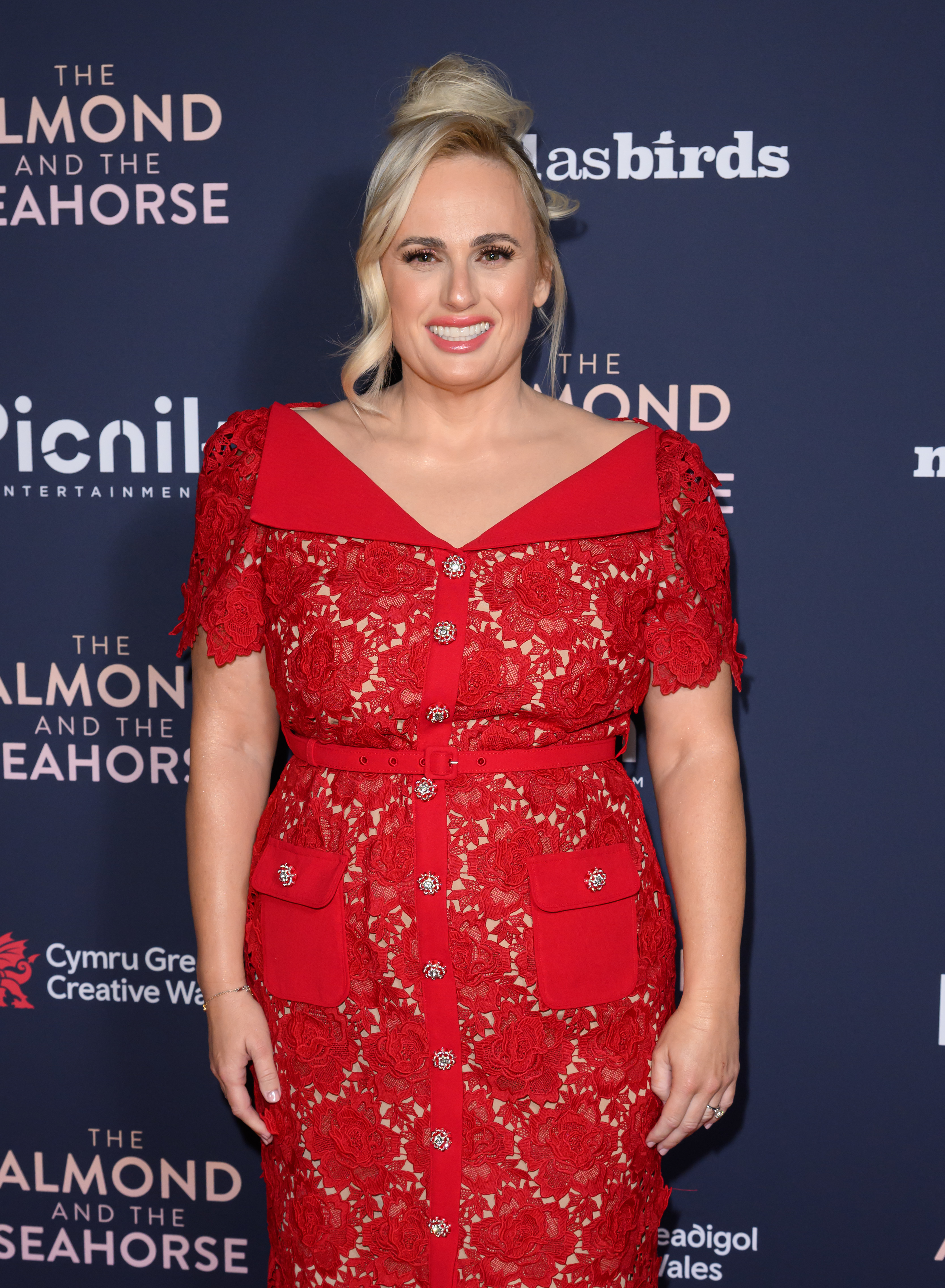 Rebel Wilson in a red lace dress with sheer sleeves at a media event
