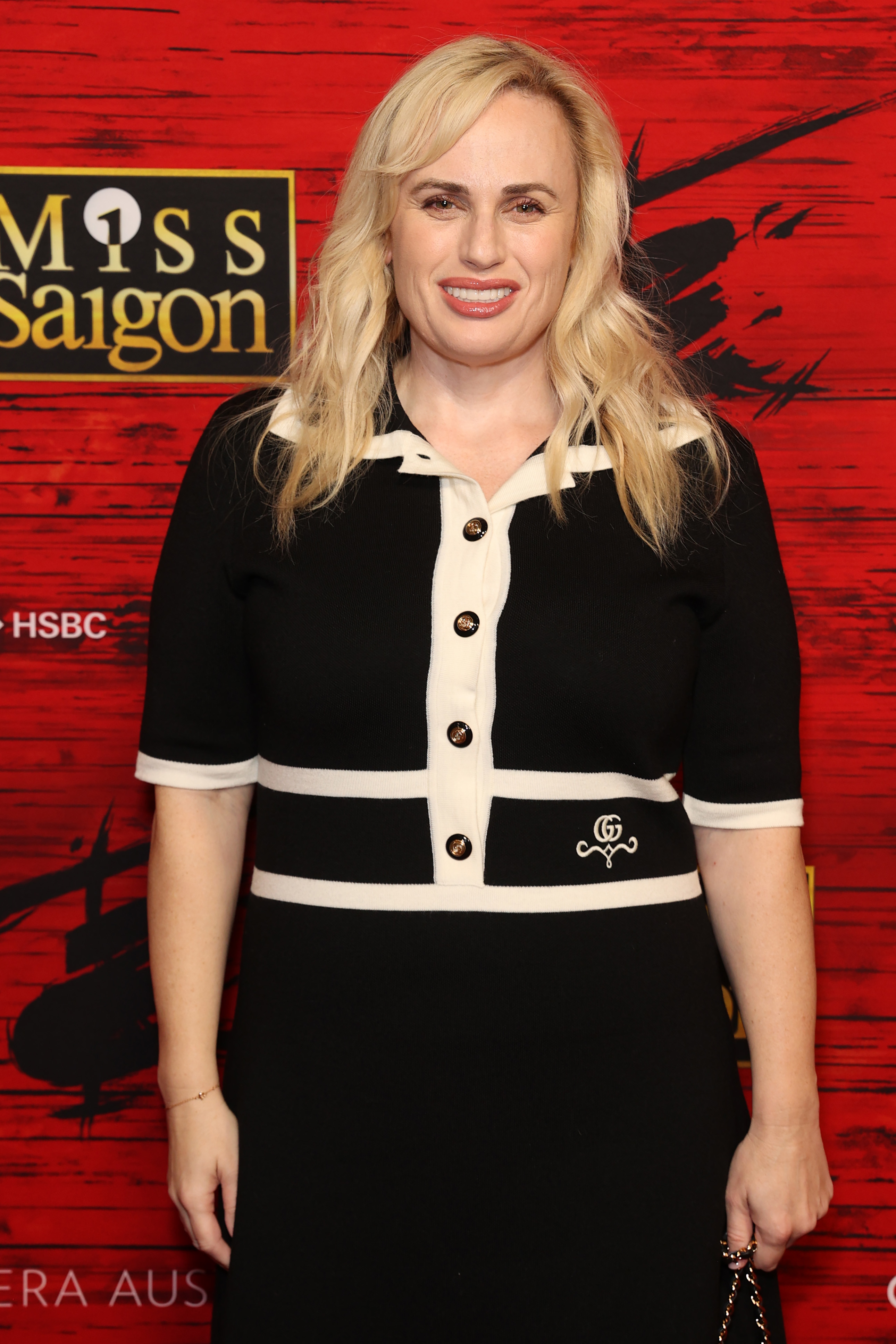 Rebel Wilson in a black dress with white trim, standing before a &#x27;Miss Saigon&#x27; backdrop