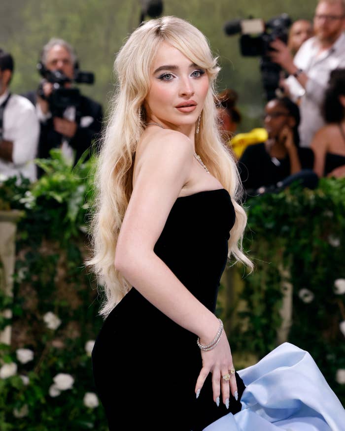 Sabrina Carpenter posing in a black strapless gown with a blue detail on the red carpet. Photographers in the background