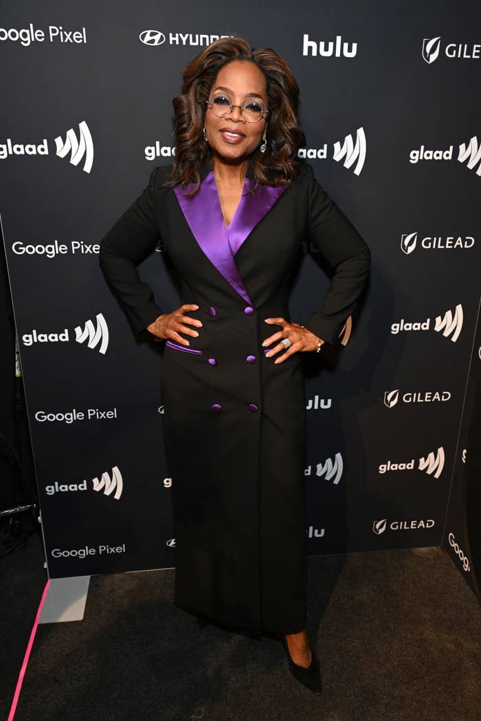 Oprah Winfrey in a black suit with purple detail, posing at an event with sponsor logos in the background