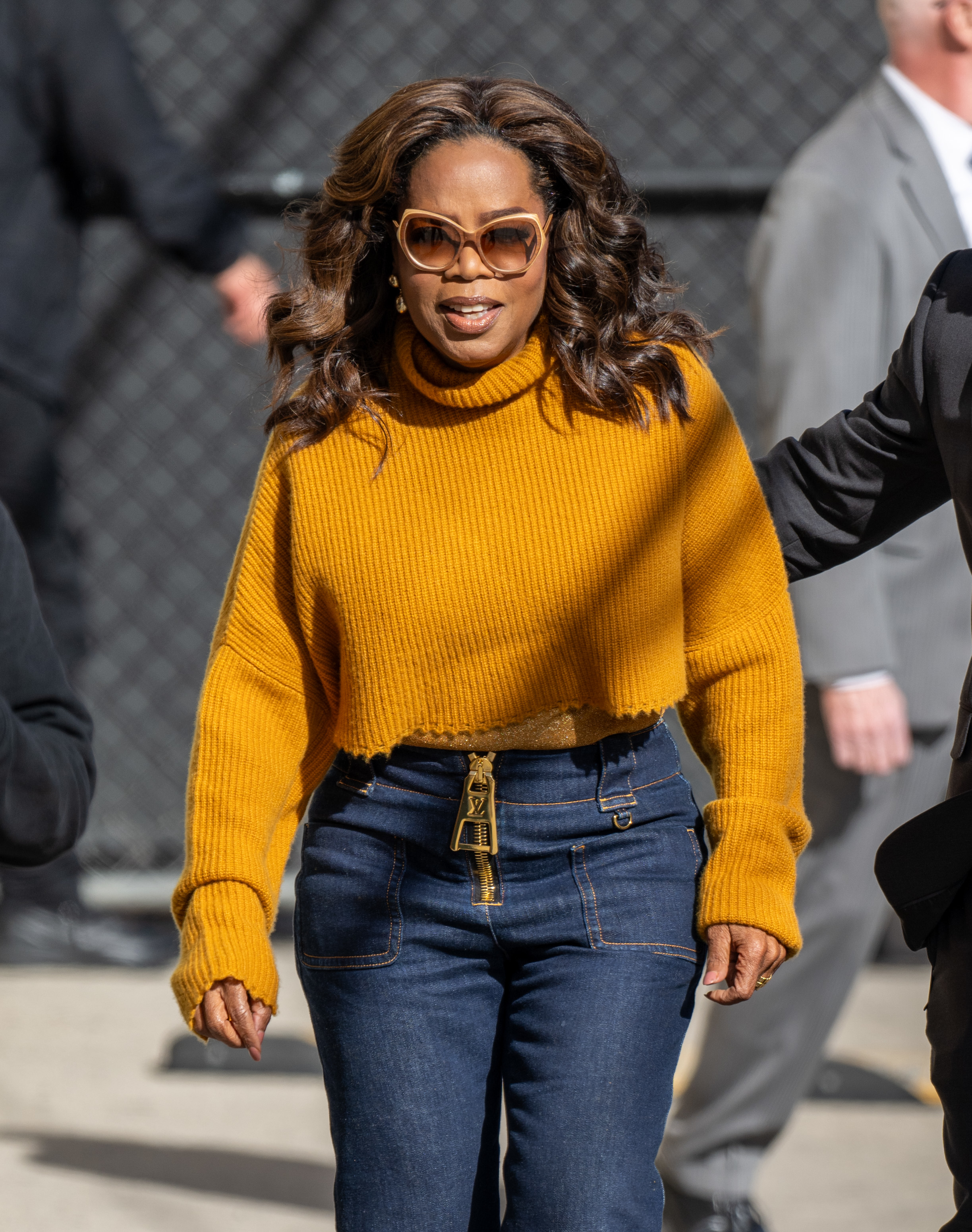 Oprah Winfrey in a yellow knit top and blue jeans, walking with sunglasses on
