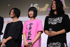 ステージに立つ3人の人物、中央の人物がピンクのTシャツを着用、マイクを持っている。