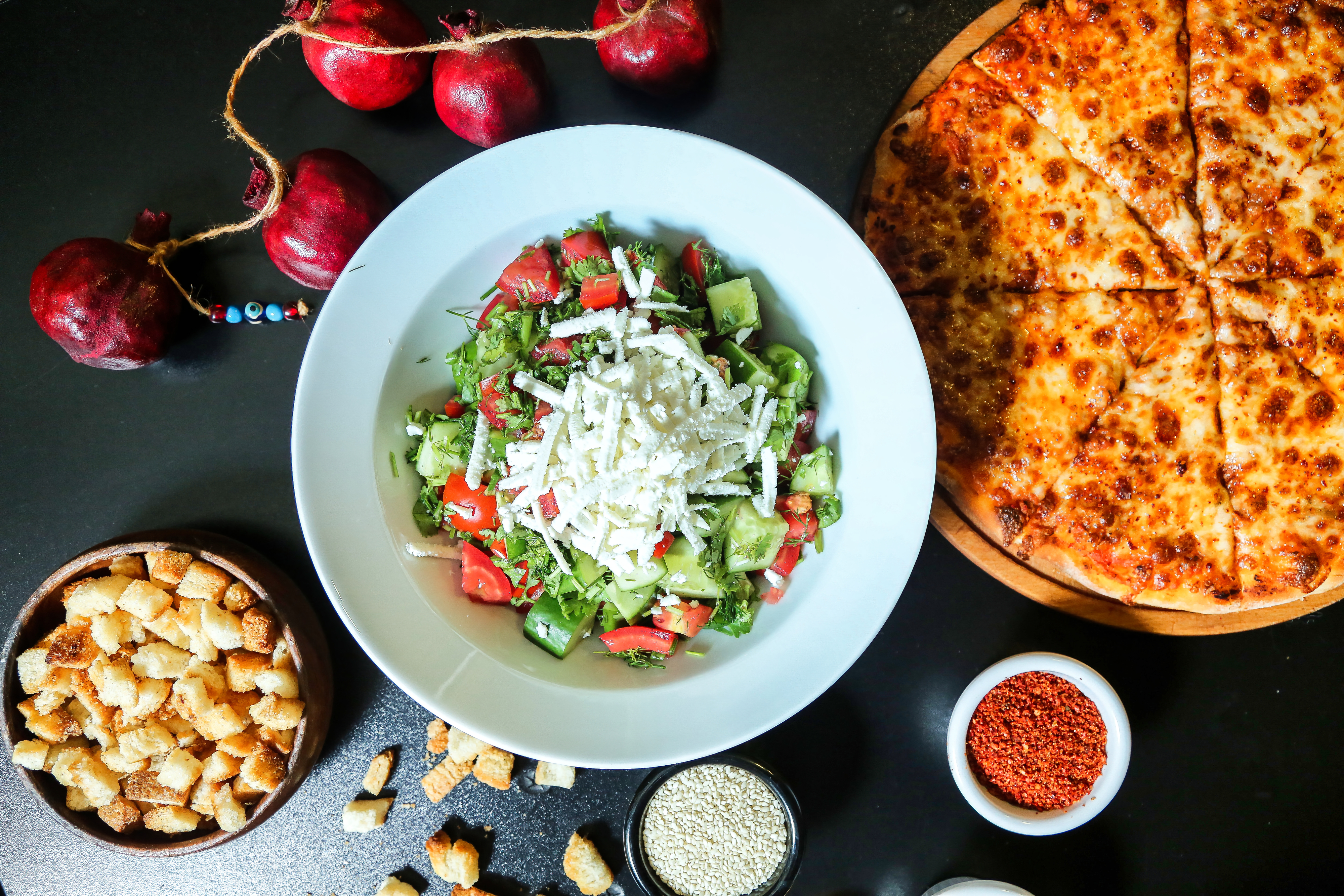 Mixed salad in a bowl near a pizza, croutons, and spices on a table