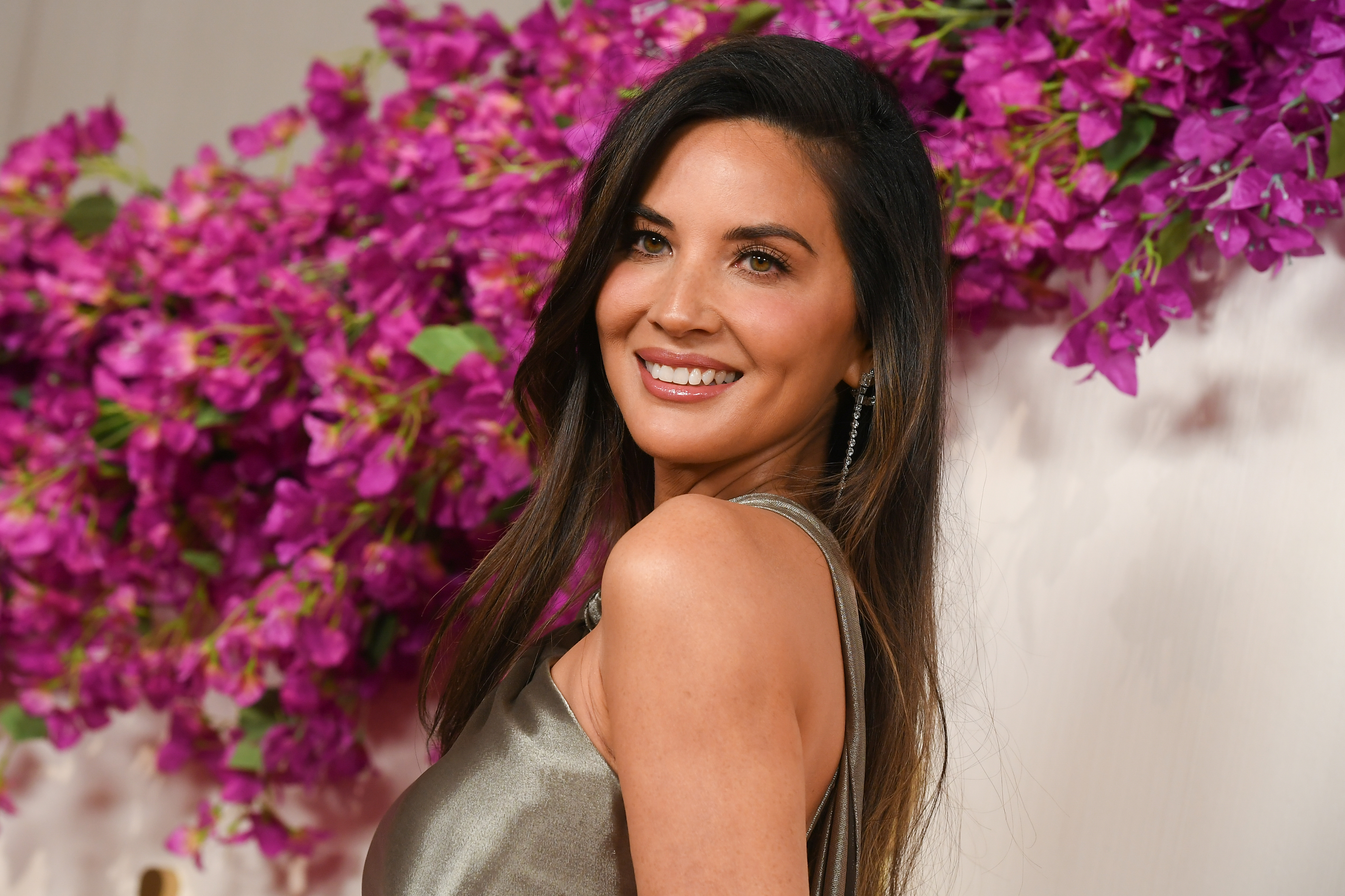 Olivia smiling on the red carpet in a sleeveless gown with flowers in the background