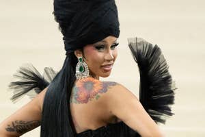 Cardi B in a black gown with an oversized headpiece and feather details, posing on the Met Gala carpet