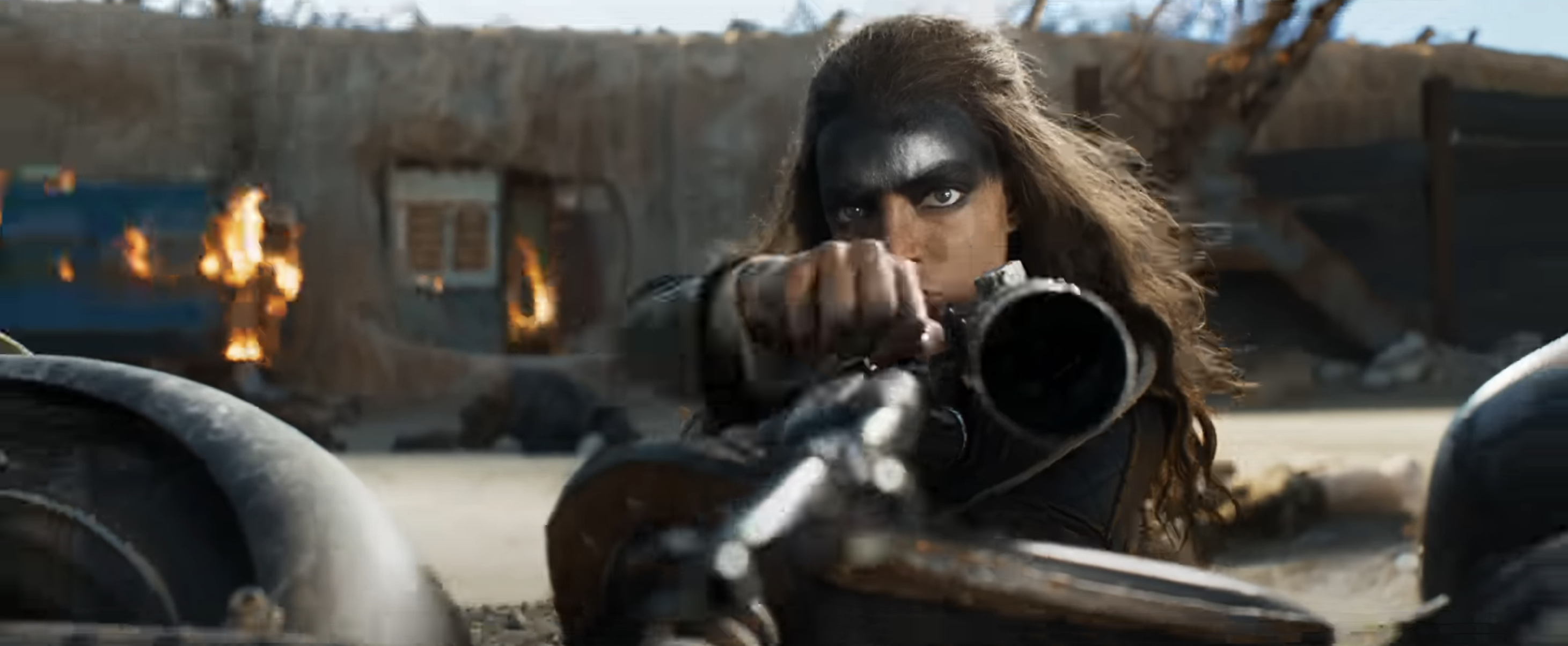 Furiosa in elaborate warrior makeup aims a crossbow, intense expression, post-apocalyptic backdrop