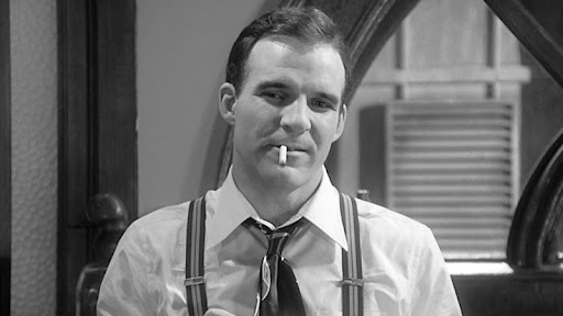 Man in a shirt and suspenders with a cigarette in his mouth, looking at the camera