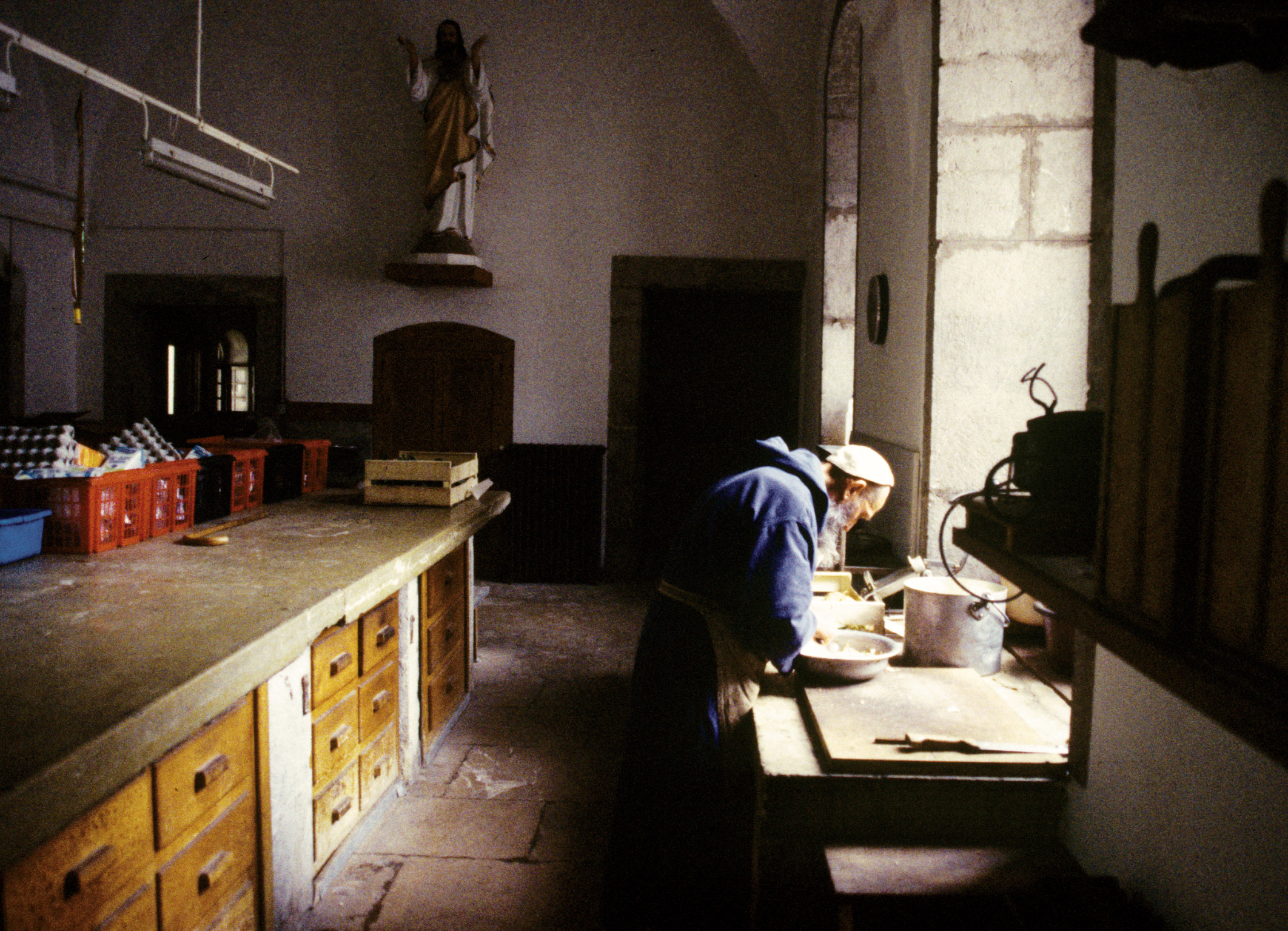 Person in traditional dress kneading dough in a rustic kitchen with religious statue in background