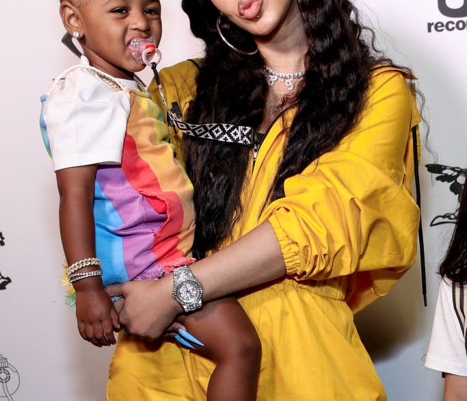 Cardi B in a draped outfit holding her daughter, who&#x27;s in a colorful dress with a yellow bow in her hair