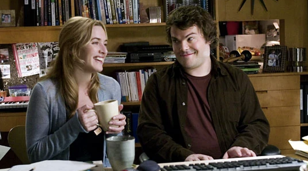 Two characters from a film sitting side by side smiling, with a bookshelf and posters in the background