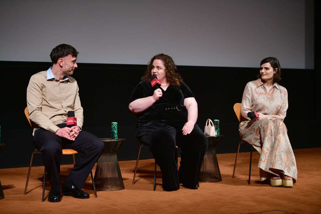 Three individuals on a stage, middle person speaks into microphone, flanked by two others, seated, attentive