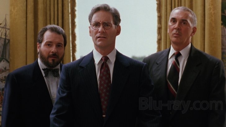 Three men in suits stand side by side; one has glasses and a bow tie, and they appear to be in a formal setting