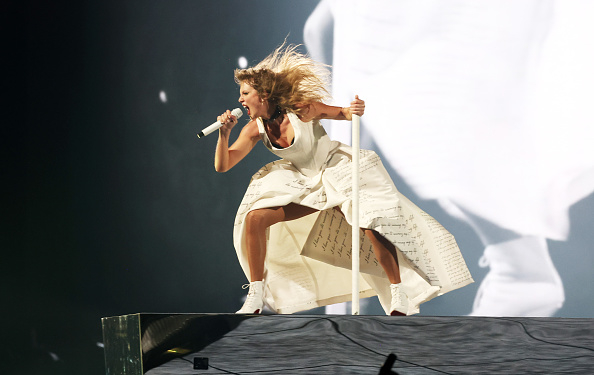 Taylor Swift performs onstage with a microphone, wearing a white dress with music note designs, in motion