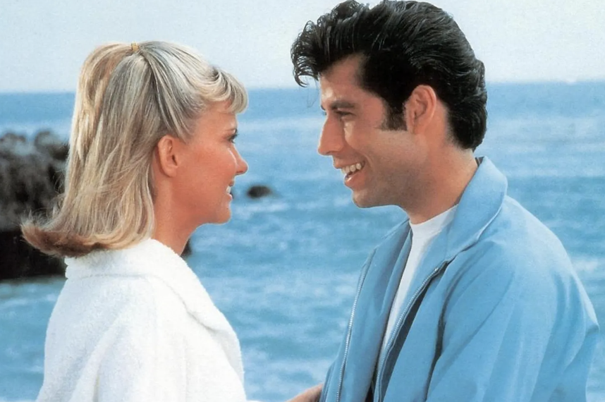 Two characters from the movie &#x27;Grease&#x27; sharing a smile, with a beach background
